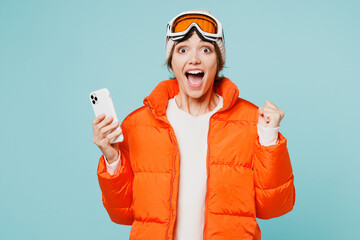 Skier winner young woman wearing warm padded windbreaker jacket hat ski goggles mask hold use mobile cell phone travel rest spend weekend winter season in mountains isolated on plain blue background. © ViDi Studio