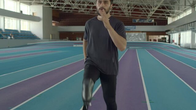 Tilt slowmo shot of determined Caucasian male runner with disability using prosthetic running blade to train on indoor stadium