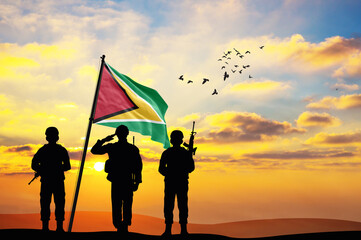 Silhouettes of soldiers with the Guyana flag stand against the background of a sunset or sunrise....