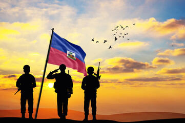 Silhouettes of soldiers with the Haiti flag stand against the background of a sunset or sunrise....