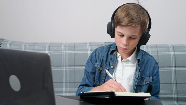 Distance learning online by video call, teen boy sitting at workplace in living room and uses a webcam to study in self-isolation at home, writes in a notebook.
