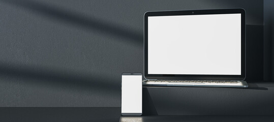 Close up of empty white laptop and cellphone on dark gray desk. Concrete wall background, shadows and pedestal or podium. Device presentation and online education concept. Mock up, 3D Rendering.