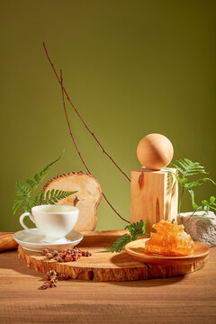 Teacups, natural raw beeswax, a wooden ball and fern leaves are displayed on a log on a wooden table with a moss green background. Front view.