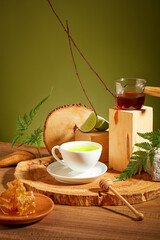 A cup of tea is placed in the center on a log, surrounded by decorations with honey, fresh lemon, a wooden platform and fern leaves. Scene for healthy tea advertisement.