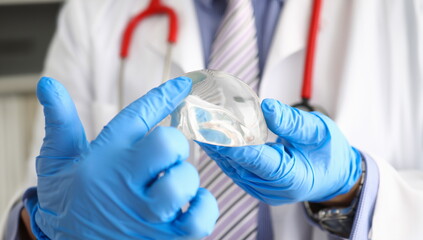 Male doctor hand in blue protective gloves hold new fashion silicone breast implant closeup....