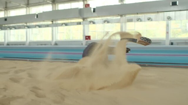 Slowmo of Caucasian male athlete with limb prosthesis doing long jump in sandy pit on indoor track
