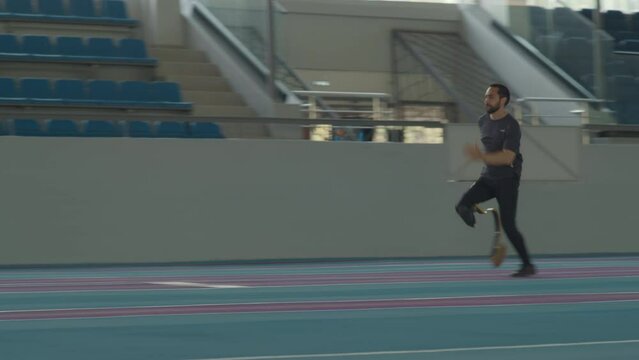 Slowmo of young Caucasian man with prosthetic limb falling off on sandy surface while practicing long jump on indoor track at stadium