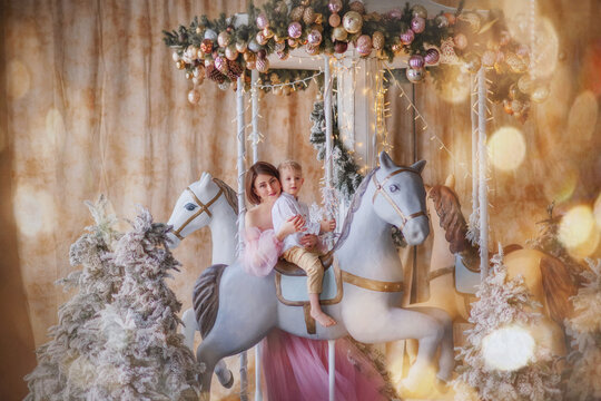 Holiday. New Year. Christmas. Carousel. Attraction. A boy in a white shirt and beige trousers sits on a horse. Mom tenderly holds her son. New Year's interior. A girl in a beautiful dress with her son