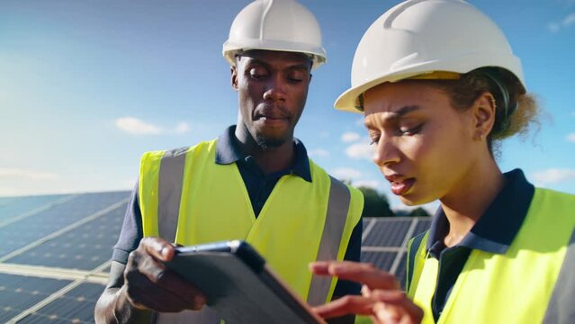 Male and female engineers wearing hard hats and hi vis safety vests with digital tablet outdoors inspecting solar panels on sustainable energy farm - shot in slow motion