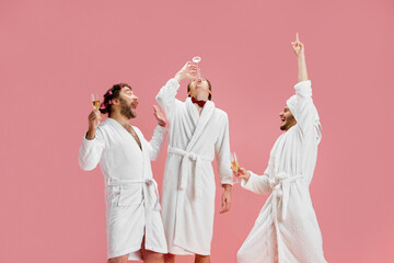 Men, friends in bathrobes and slippers drinking champagne against pink background. Preparation for...