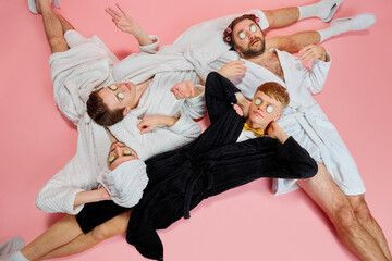 Young men, friends with cucumber eye mask lying on floor in bathrobes against pink background....