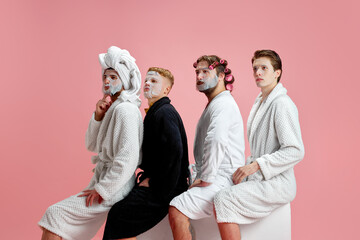 Handsome men, friends in bathrobes standing in face masks, taking care after skin against pink background. Concept of leisure activity, fun, bachelor party, friendship, spa, cosmetology