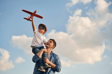 Orange colored toy plane in hands. Father and little son are playing and having fun outdoors - obrazy, fototapety, plakaty