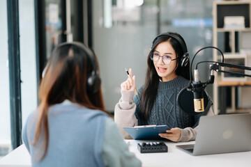 Young woman and Asian woman wearing headphones and doing a live podcast for their channel,...