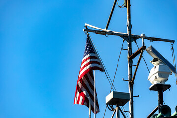 Photo of the large American flag, flying on a flagpole of an American ship under a blue sky, from...