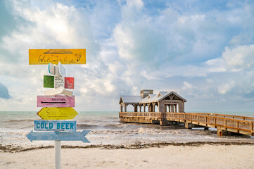A colourful, wooden signpost, on a beach, with a wooden jetty in the background.