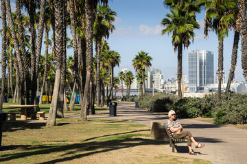 Elderly woman sits on a bench in the Coronado Tidelands park between palms and the San Diego...