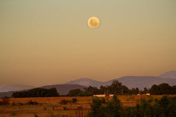 Full moon over a field, trees and mountains against an orange and blue glowing sky at sunset.