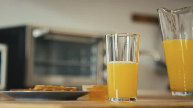 Side stab shot of hand filling glass with orange juice from jug and taking glass from table in kitchen