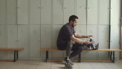Young Caucasian man with prosthetic limb arriving in locker room, putting sports bag on wooden bench and starting taking out necessities - Powered by Adobe
