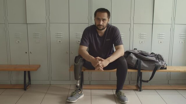 Portrait of serious Caucasian male athlete with prosthetic leg sitting on wooden bench in modern locker room and posing for camera