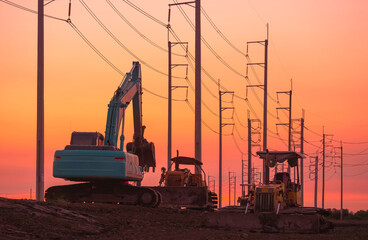 Silhouette of excavator and 2 bulldozer tractors parked on country road with row of electric poles against orange sunset sky background in the evening, perspective side view