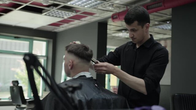 A male brunette hairdresser cuts a haircut for a handsome young man in a barbershop, general plan. Modern and fashionable haircut. Professional barbershop. Slow motion