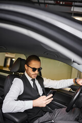 appealing red haired man with beard and sunglasses looking at phone behind steering wheel, business