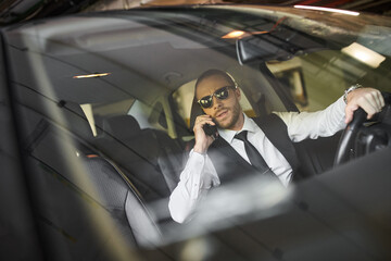 attractive bearded man with ponytail and sunglasses talking by phone behind steering wheel, business
