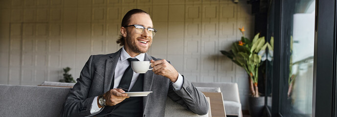 handsome cheerful businessman in elegant suit smiling sitting on sofa and drinking tea, banner