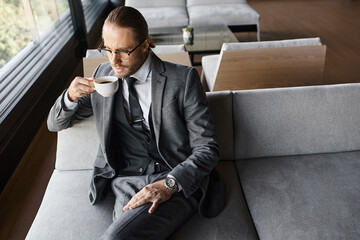 attractive elegant man in smart gray suit with glasses drinking warm tea on sofa, business concept