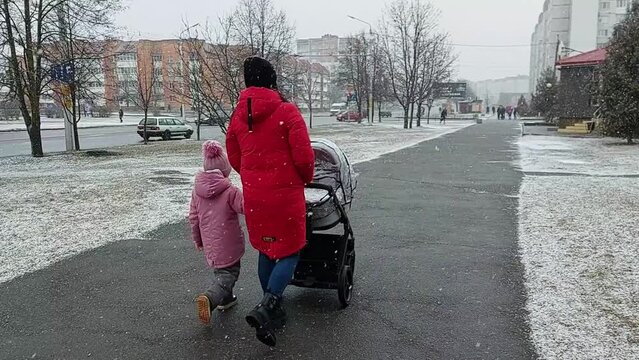 A mother in a warm red coat with her daughter and a stroller are walking around the city in winter weather with flakes of wet snow. Early winter.