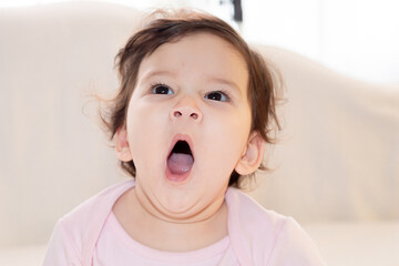 Close-up cute infant girl yawning with big wide mouth to camera sitting in bed alone feeling sleepy...