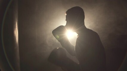 Silhouette of young man doing boxing training at the gym, hitting the punching bag wearing boxing gloves