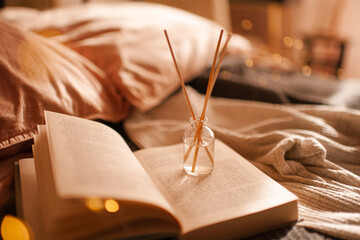 Home liquid fragrance in glass bottle with bamboo sticks on paper book in bed over glow lights close up. Aromatherapy.