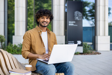 Portrait of a young smiling Indian man sitting on a bench outside with a laptop on his lap, working...