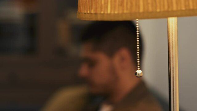 Side stab closeup of blurred bearded man sitting on sofa and turning on floor lamp by pulling chain in modern house