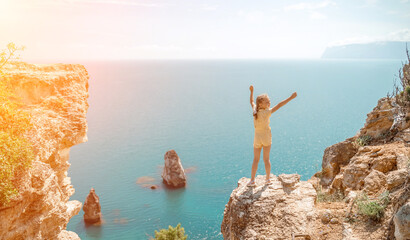 Happy girl stands on a rock high above the sea, wearing a yellow jumpsuit and sporting braided...