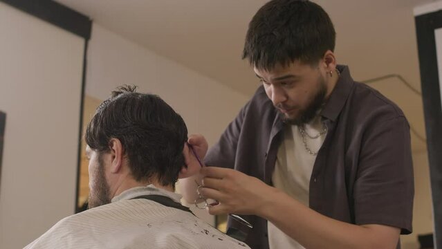 Young professional barber cutting hair of man with scissors and comb during workday in barbershop