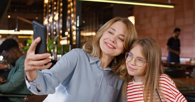 Loving mother making photo with her cute daughter on phone. Caucasian family smiling on phone camera while making selfie. Enjoying time together. People resting at cafe. Family day.