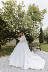 Obraz na płótnie Canvas Curly brunette bride in a lace dress with an open bust, in a lush veil holds a bouquet and poses against the background of green trees. Spring wedding