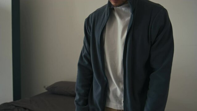 Midsection cropped shot of unrecognizable man putting on dark sweat jacket on white t-shirt and zipping it up while dressing up in bedroom