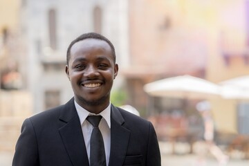 African businessman in a black suit stands in front of a small store or SME with a smile