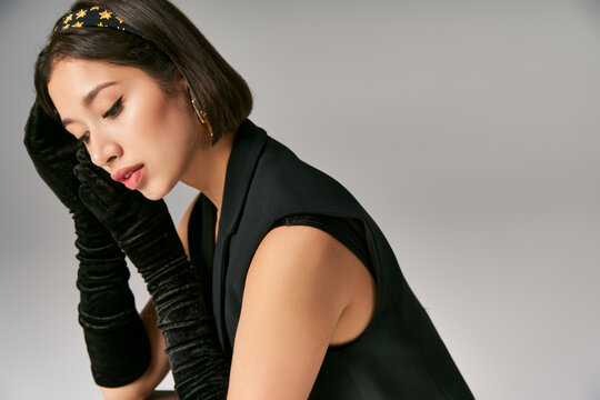 attractive and dreamy asian woman in elegant attire with gloves posing on grey background