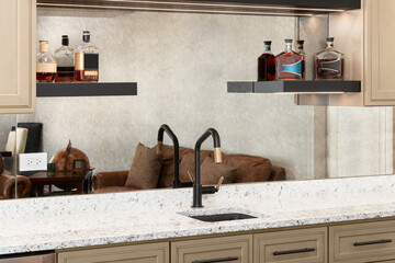 A wet bar with brown cabinets, a white countertop, bottles sitting on floating shelves, and a black...