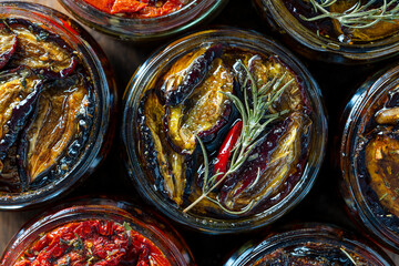 Sun-dried red tomatoes and plums with garlic, green rosemary, red chili pepper, olive oil and spices in a glass jars on a wooden table. Rustic style, top view, closeup - obrazy, fototapety, plakaty