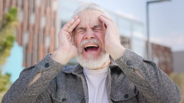 Senior male with gray beard holding head with hands and screaming from pain while standing in urban park. Portrait of stylish pensioner in coat suffering from dreadful headache while standing outside.