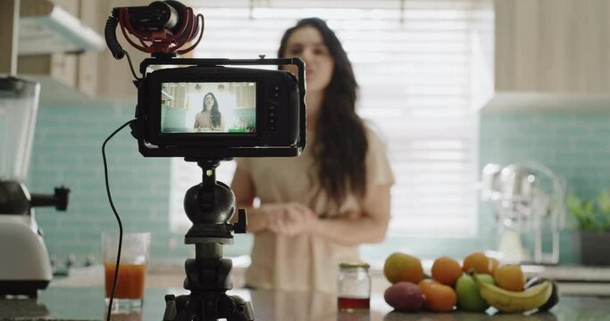 Woman, camera and live streaming in kitchen, blog and tutorial or teaching at home on broadcast. Female influencer, webinar and vlog or technology for video recording, juice and fruit for nutrition