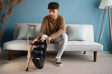 A young man with an ankle injury wearing a rigid ankle orthosis sits on the sofa with crutches at...