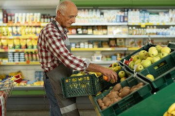 Old gray-haired man in supermarket as shop assistant holding boxes with apples and putting on...